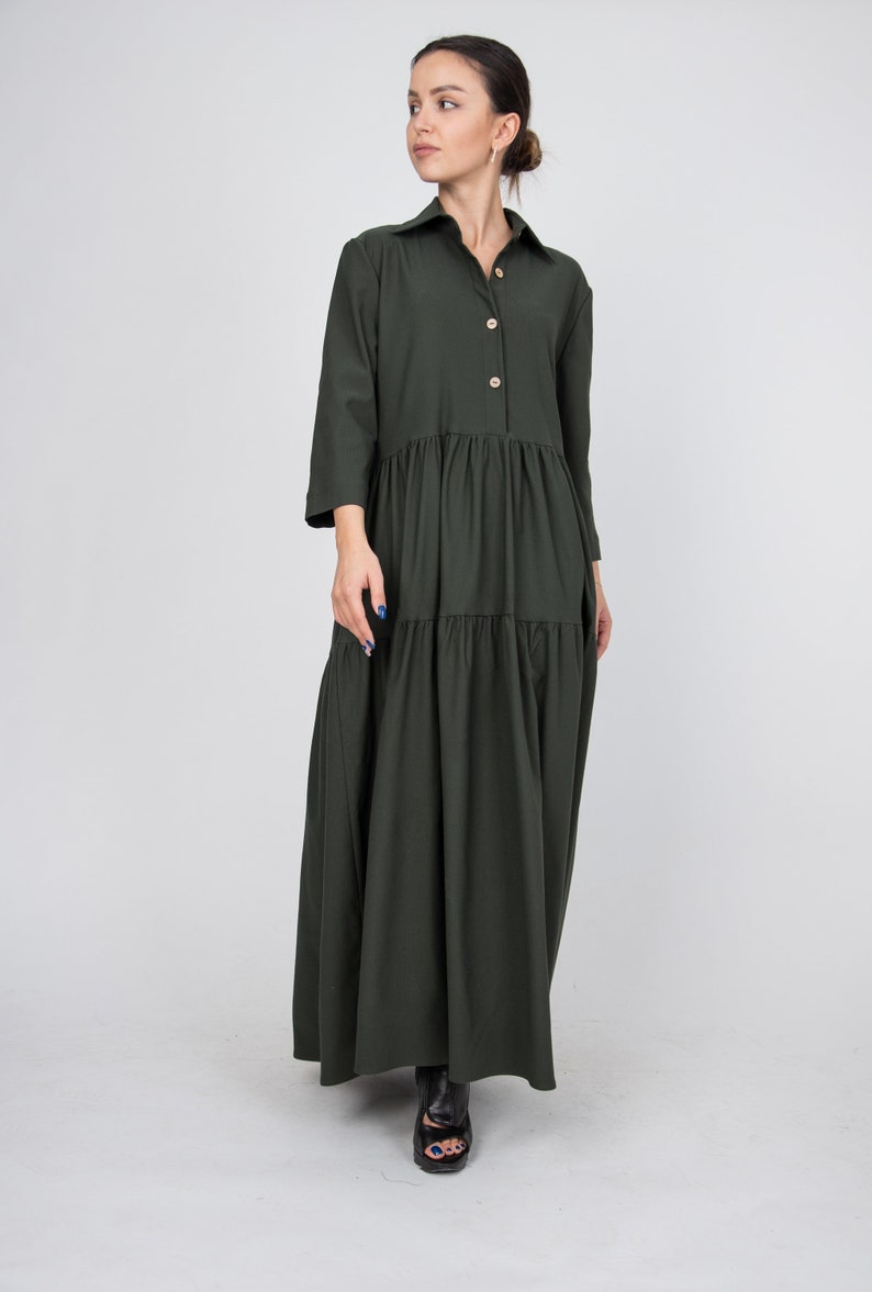 May include: A long, dark green, tiered dress with a button-up collar and long sleeves. The dress has a relaxed fit and falls to the ankles.