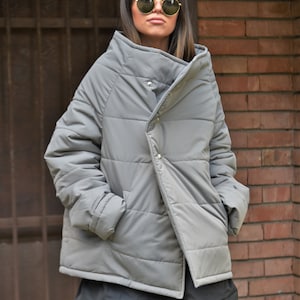 Peut inclure: Une veste matelass&eacute;e grise avec un design asym&eacute;trique unique et un col montant. La veste a une fermeture &agrave; bouton et une texture matelass&eacute;e.