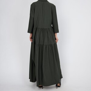 May include: A long, dark green, tiered dress with a collared neckline and long sleeves. The dress is made of a soft, flowing fabric and has a relaxed fit.