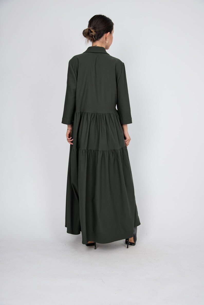 May include: A long, dark green, tiered dress with a collared neckline and long sleeves. The dress is made of a soft, flowing fabric and has a relaxed fit.