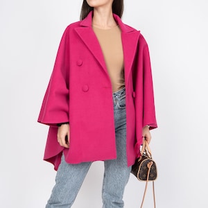 May include: A woman wearing a pink double-breasted coat with a brown top and blue jeans. She is holding a brown leather handbag with a long strap.