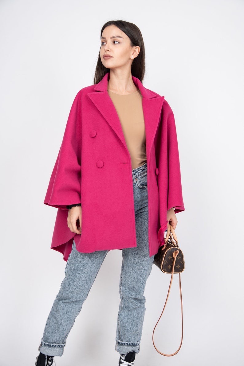 May include: A woman wearing a pink double-breasted coat with a brown top and blue jeans. She is holding a brown leather handbag with a long strap.