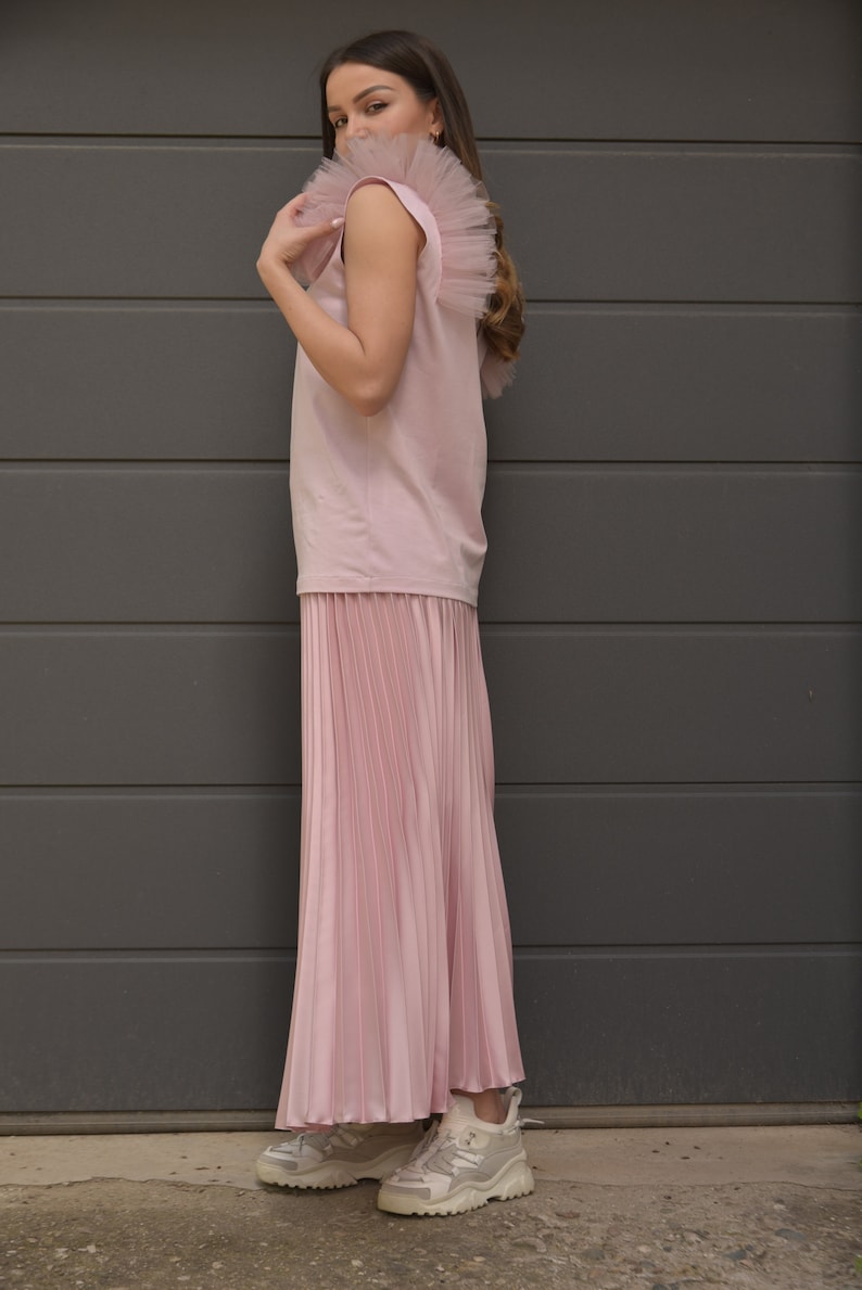 May include: A pink sleeveless top with a tulle ruffle on the shoulder and a pleated pink skirt. The person is wearing white sneakers.
