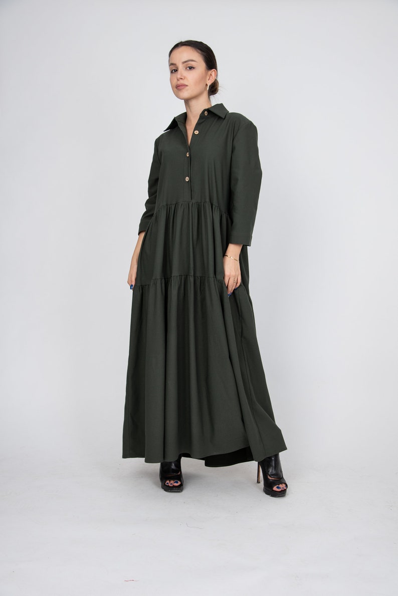May include: A long, tiered, dark green dress with a button-up front and long sleeves. The dress is made of a soft, flowing fabric and has a relaxed fit.