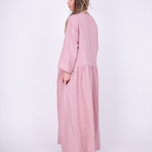 May include: A long, flowing pink linen dress with a loose fit and three-quarter sleeves. The dress has a gathered waist and a simple, round neckline.
