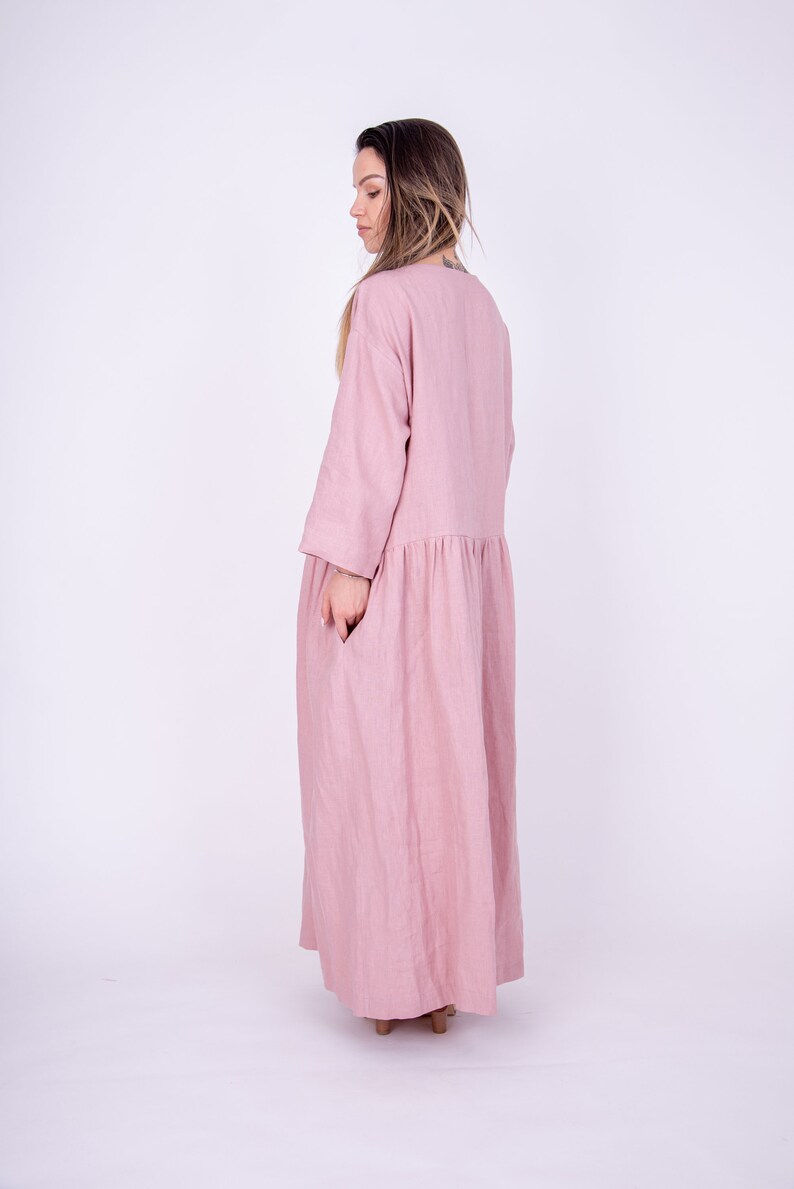 May include: A long, flowing pink linen dress with a loose fit and three-quarter sleeves. The dress has a gathered waist and a simple, round neckline.