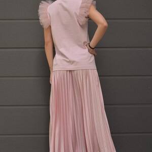 May include: A pink t-shirt with tulle ruffle sleeves and a pleated pink skirt. The model is wearing white sneakers.