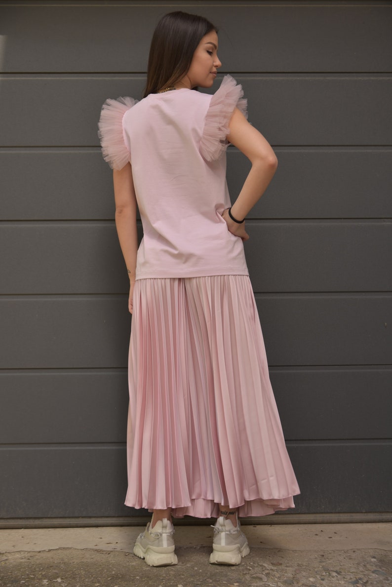 May include: A pink t-shirt with tulle ruffle sleeves and a pleated pink skirt. The model is wearing white sneakers.