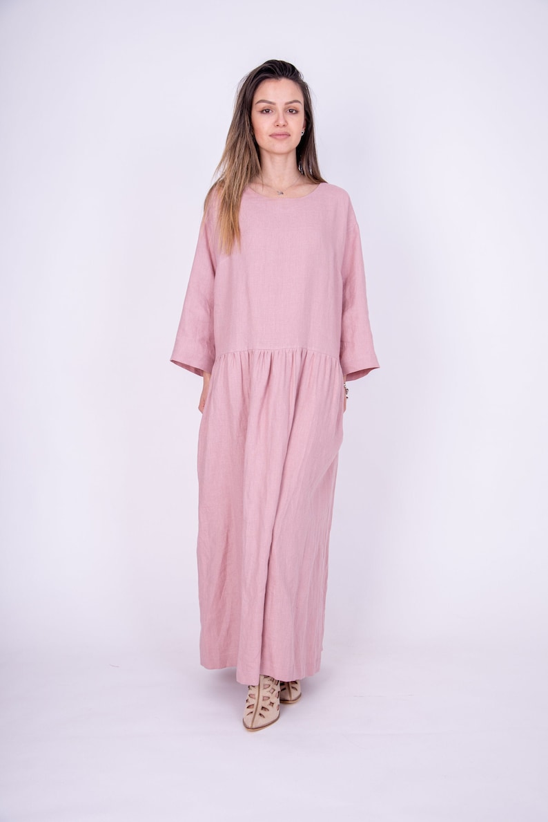 May include: A long, pink linen dress with a loose fit and 3/4 sleeves. The dress has a gathered waist and a simple, round neckline.