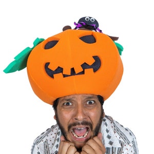 May include: A man wearing a bright orange pumpkin hat with a black spider on top. He is looking at the camera with a scared expression and his hands are covering his face.