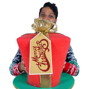 Peut inclure: Un grand cadeau de No&euml;l rouge avec un n&oelig;ud dor&eacute; et une &eacute;tiquette avec l'inscription "Merry Christmas". Le cadeau repose sur un bord vert. La personne qui tient le cadeau porte un pull de No&euml;l.