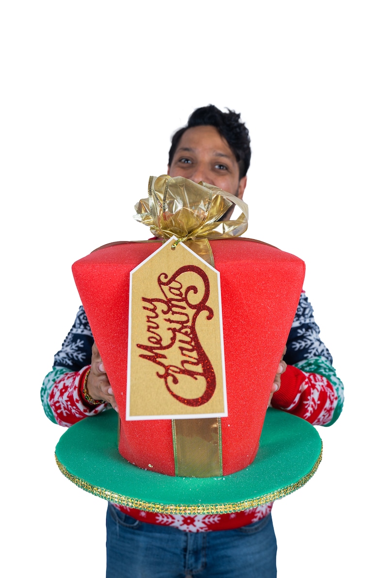 Peut inclure: Un grand cadeau de No&euml;l rouge avec un n&oelig;ud dor&eacute; et une &eacute;tiquette avec l'inscription "Merry Christmas". Le cadeau repose sur un bord vert. La personne qui tient le cadeau porte un pull de No&euml;l.