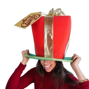 Peut inclure: Un chapeau de No&euml;l fantaisie en forme de bo&icirc;te cadeau rouge avec un ruban dor&eacute; et une &eacute;tiquette avec l'inscription "Merry Christmas". Le chapeau a un bord vert et est tenu par une personne.