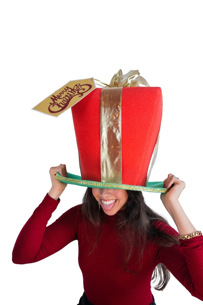 Peut inclure: Un chapeau de No&euml;l fantaisie en forme de bo&icirc;te cadeau rouge avec un ruban dor&eacute; et une &eacute;tiquette avec l'inscription "Merry Christmas". Le chapeau a un bord vert et est tenu par une personne.