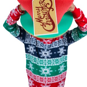 Peut inclure: Une personne portant un pull de No&euml;l et tenant une grande bo&icirc;te cadeau rouge avec une &eacute;tiquette dor&eacute;e qui dit "Merry Christmas". Le pull a un motif de flocons de neige en rouge, vert et bleu marine.