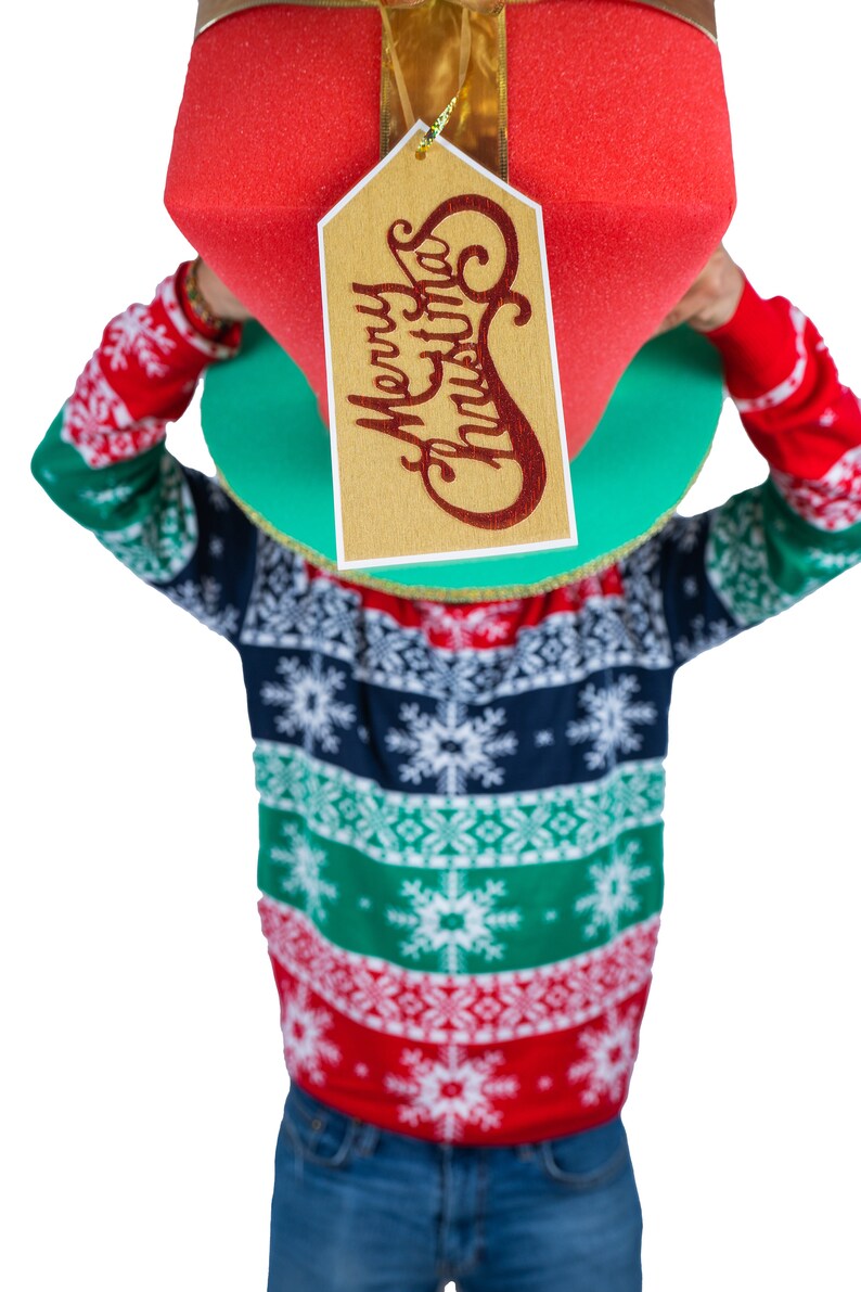Peut inclure: Une personne portant un pull de No&euml;l et tenant une grande bo&icirc;te cadeau rouge avec une &eacute;tiquette dor&eacute;e qui dit "Merry Christmas". Le pull a un motif de flocons de neige en rouge, vert et bleu marine.