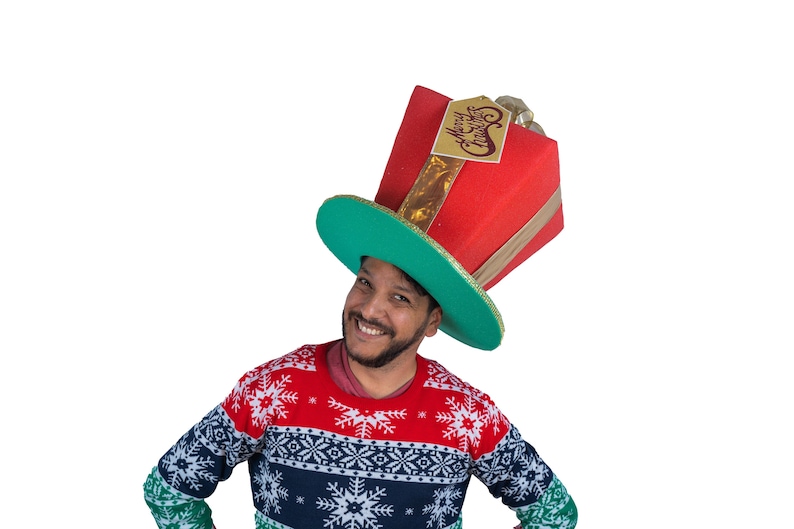 Peut inclure: Un chapeau de No&euml;l fantaisie en forme de bo&icirc;te cadeau rouge avec un bord vert et des accents dor&eacute;s. Le chapeau porte une &eacute;tiquette avec l'inscription "Merry Christmas". La personne porte un pull &agrave; motifs flocons de neige rouge, blanc et bleu.