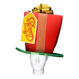 Peut inclure: Un chapeau de No&euml;l fantaisie en forme de bo&icirc;te cadeau rouge, orn&eacute; d'un n&oelig;ud dor&eacute; et d'un bord vert. Le chapeau porte une &eacute;tiquette dor&eacute;e avec l'inscription "Merry Christmas".