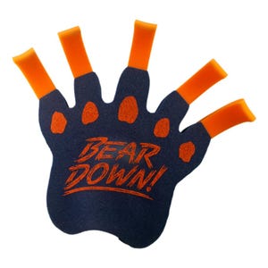 Pode incluir: Uma mão de espuma azul-escura em forma de pata de urso com cinco pontas de dedos cor de laranja. A palma apresenta detalhes de almofadas cor de laranja brilhantes e o texto BEAR DOWN! impresso em laranja no centro do acessório de espuma.