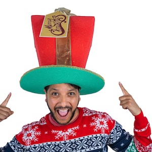 Peut inclure: Une personne porte un chapeau de No&euml;l fantaisie en forme de bo&icirc;te cadeau. Le chapeau est rouge avec un bord vert et une &eacute;tiquette dor&eacute;e qui dit "Merry Christmas". La personne porte &eacute;galement un pull de No&euml;l rouge, blanc et bleu.