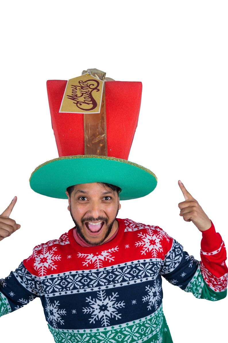 Peut inclure: Une personne porte un chapeau de No&euml;l fantaisie en forme de bo&icirc;te cadeau. Le chapeau est rouge avec un bord vert et une &eacute;tiquette dor&eacute;e qui dit "Merry Christmas". La personne porte &eacute;galement un pull de No&euml;l rouge, blanc et bleu.