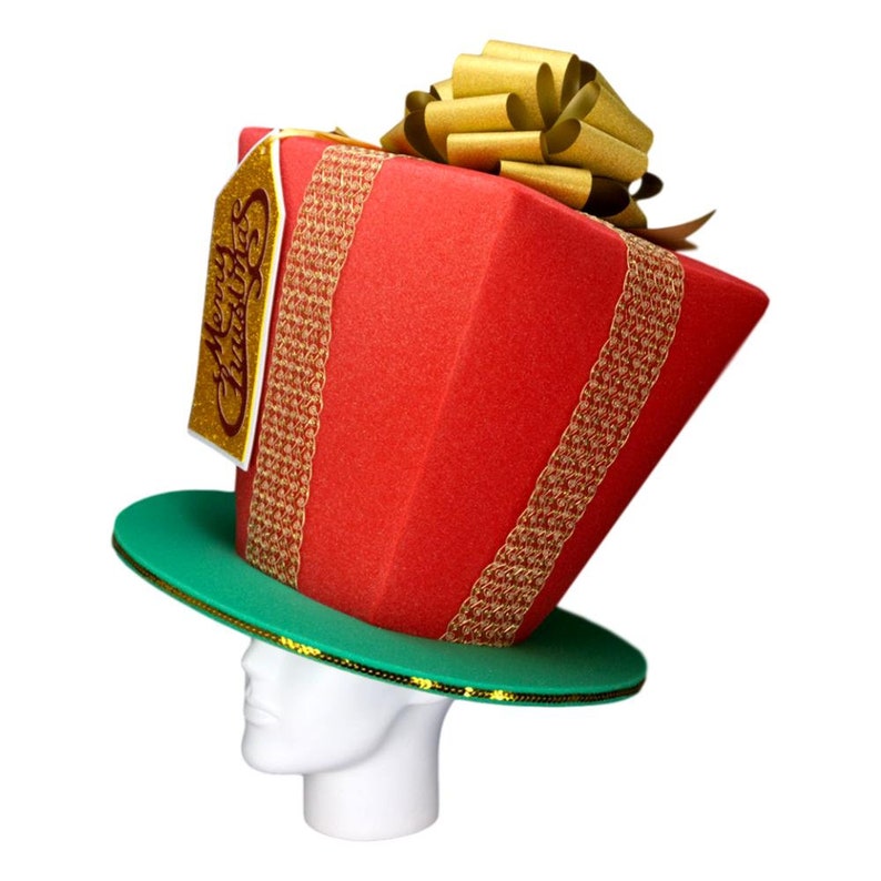 Peut inclure: Un chapeau de No&euml;l fantaisie en forme de bo&icirc;te cadeau rouge avec des rubans et des bordures dor&eacute;s. Le chapeau repose sur un bord vert et comporte une &eacute;tiquette dor&eacute;e avec l'inscription "Merry Christmas".