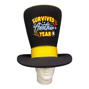 Op de afbeelding: Een zwarte hoge hoed met een gele band en rand. De hoed heeft de tekst "SURVIVED Another YEAR" in kleurrijke letters. De hoed wordt gedragen door een mannequinhoofd.