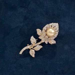 May include: A gold rose brooch with a pearl centre and diamond accents. The brooch is displayed on a dark blue velvet background.