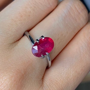 以下が含まれることがあります： 大きな楕円形のルビーの宝石がついたシルバーリング。ルビーは濃い赤色で、シンプルでエレガントなセッティングにセットされています。