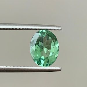 May include: A green oval gemstone held by tweezers. The gemstone is faceted and has a light green color.