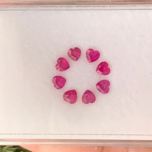 May include: A small white box with a clear lid containing a circle of ten pink heart-shaped gemstones.