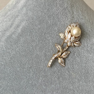 May include: A gold tone brooch in the shape of a rose with a single pearl in the centre. The rose is made of clear rhinestones.