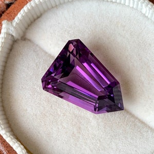 May include: A deep purple amethyst gemstone with a faceted cut, displayed in a white velvet ring box.
