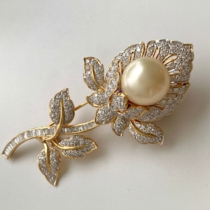 May include: A gold-tone brooch in the shape of a flower with a large pearl in the centre. The flower is adorned with small, clear rhinestones.