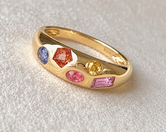 May include: A gold band ring featuring five multi-colored gemstones set into the metal. The stones include a round blue sapphire, a pentagon-shaped orange stone, a round yellow stone, an oval pink stone, and a rectangular pink stone.