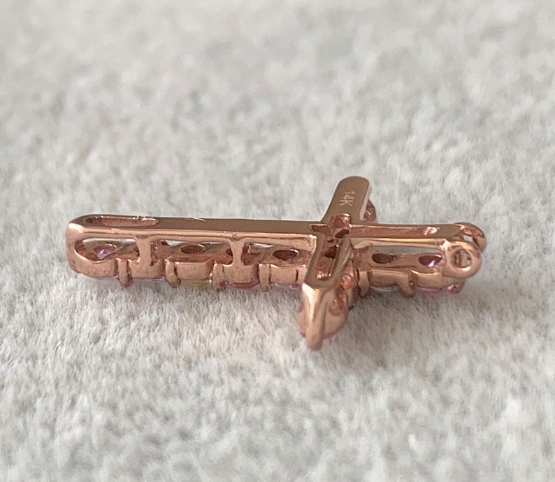 May include: A rose gold cross pendant with small pink gemstones set around the edges. The pendant is marked "14K" on the back.