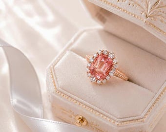 May include: A ring featuring a rectangular, peach-colored center gemstone surrounded by a halo of small, round, clear stones. The band is crafted from a metallic material with a textured, beaded design, presented in a light-colored jewelry box.