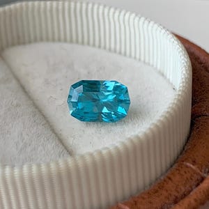 Puede incluir: Una gema azul vibrante con un corte rectangular, expuesta en una caja de anillo de terciopelo blanco.