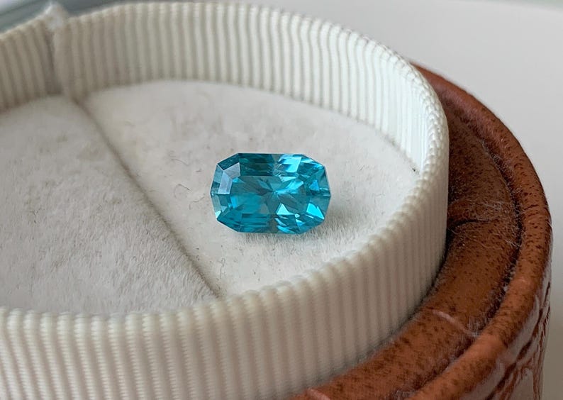 Puede incluir: Una gema azul vibrante con un corte rectangular, expuesta en una caja de anillo de terciopelo blanco.