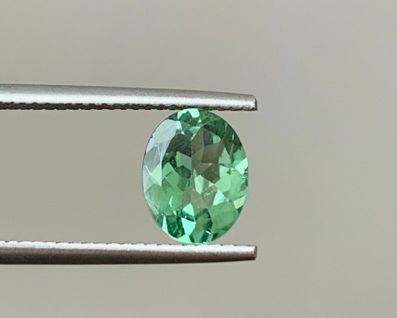 May include: A green oval gemstone held by tweezers. The gemstone is faceted and has a light green color.