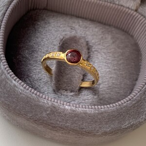May include: A gold ring with a single, round, dark pink gemstone surrounded by a row of small, clear gemstones.