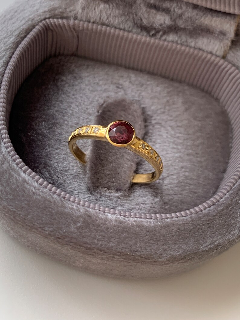 May include: A gold ring with a single, round, dark pink gemstone surrounded by a row of small, clear gemstones.