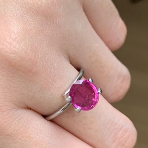 Pode incluir: Um anel de prata com uma grande pedra rosa vibrante de forma redonda. A pedra é presa por quatro garras, e a banda curva-se elegantemente. Uma joia marcante e elegante.