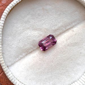 May include: A vibrant purple gemstone with a rectangular emerald cut, displayed on a white velvet background.