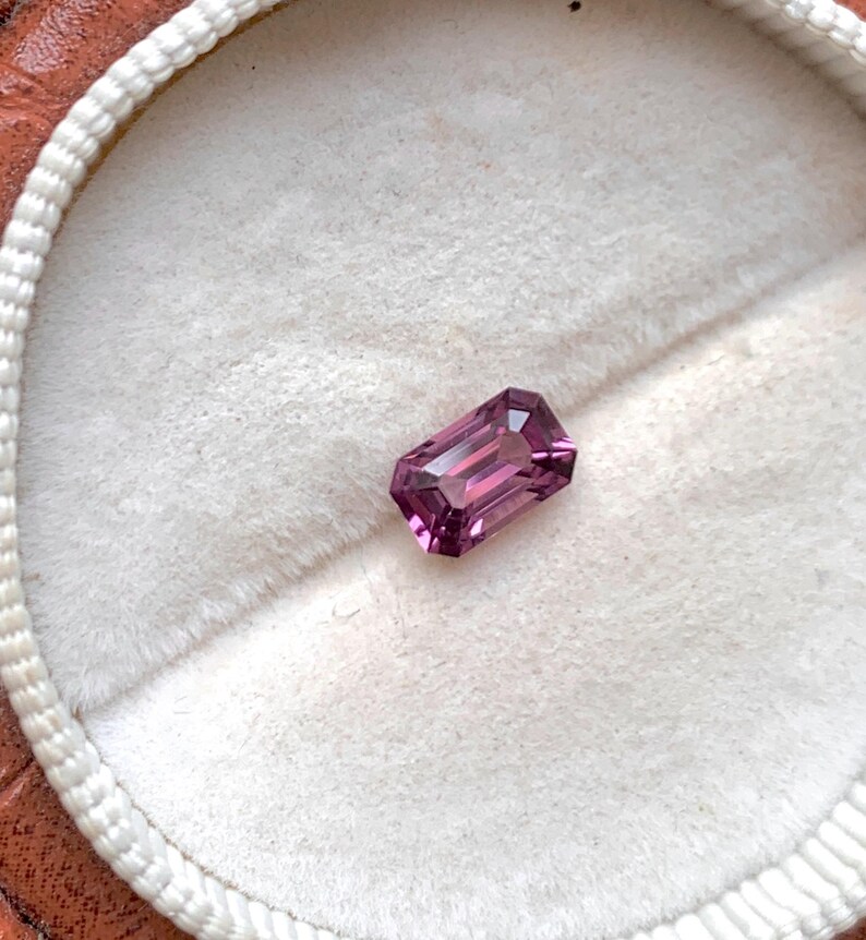 May include: A vibrant purple gemstone with a rectangular emerald cut, displayed on a white velvet background.