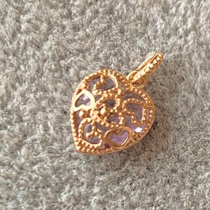 K&ouml;nnte beinhalten: Ein goldfarbener herzf&ouml;rmiger Anh&auml;nger mit einem filigranen Design und einem hellvioletten Zentrum.