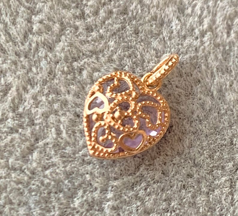 K&ouml;nnte beinhalten: Ein goldfarbener herzf&ouml;rmiger Anh&auml;nger mit einem filigranen Design und einem hellvioletten Zentrum.