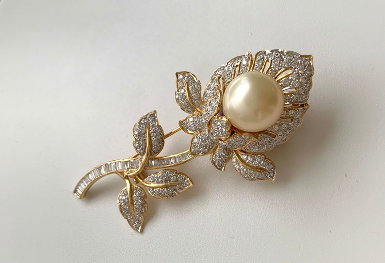 May include: A gold-tone brooch in the shape of a flower with a large pearl in the centre. The flower is adorned with small, clear rhinestones.