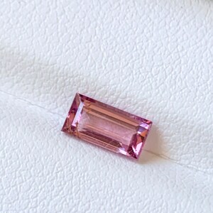 Op de afbeelding: Een rechthoekige, roze-paarse edelsteen, geplaatst op een witte, getextureerde achtergrond. De steen is een baguette-slijpsel, met parallelle zijden en een trapsgewijze kroon. Ongeveer 1,5 cm lang.