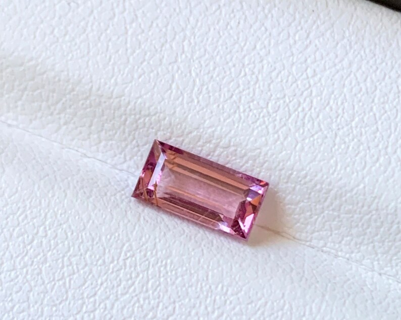Op de afbeelding: Een rechthoekige, roze-paarse edelsteen, geplaatst op een witte, getextureerde achtergrond. De steen is een baguette-slijpsel, met parallelle zijden en een trapsgewijze kroon. Ongeveer 1,5 cm lang.